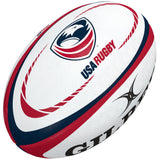 The Gilbert USA Replica Rugby Ball features red and blue curved stripes, the USA Rugby logo, and a partial black Gilbert brand logo—a perfect gift for rugby fans or collectors.