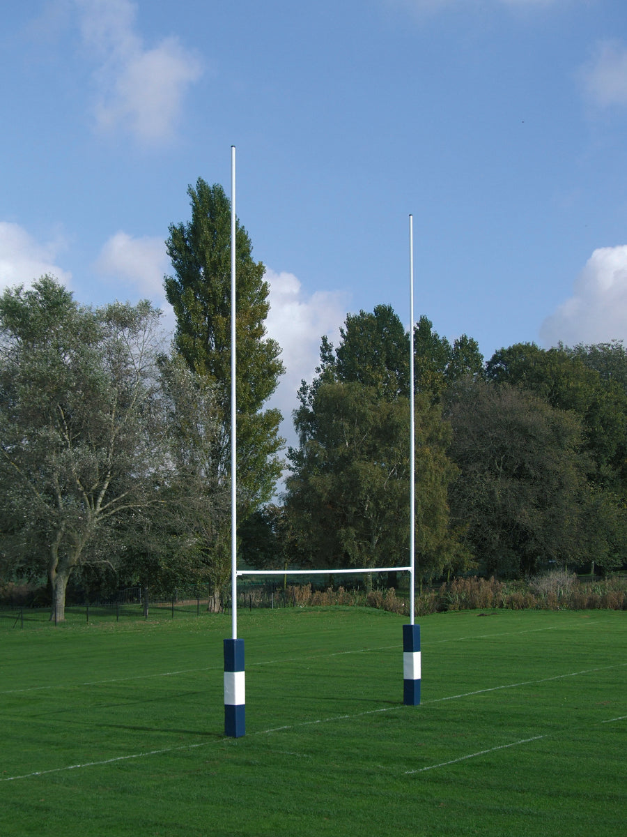 The Harrod Sport No. 2 Steel Rugby Posts - 10M Hinged stand tall with blue and white padded bases on a green field, surrounded by trees under a blue sky—rugged steel construction ensures lasting performance.