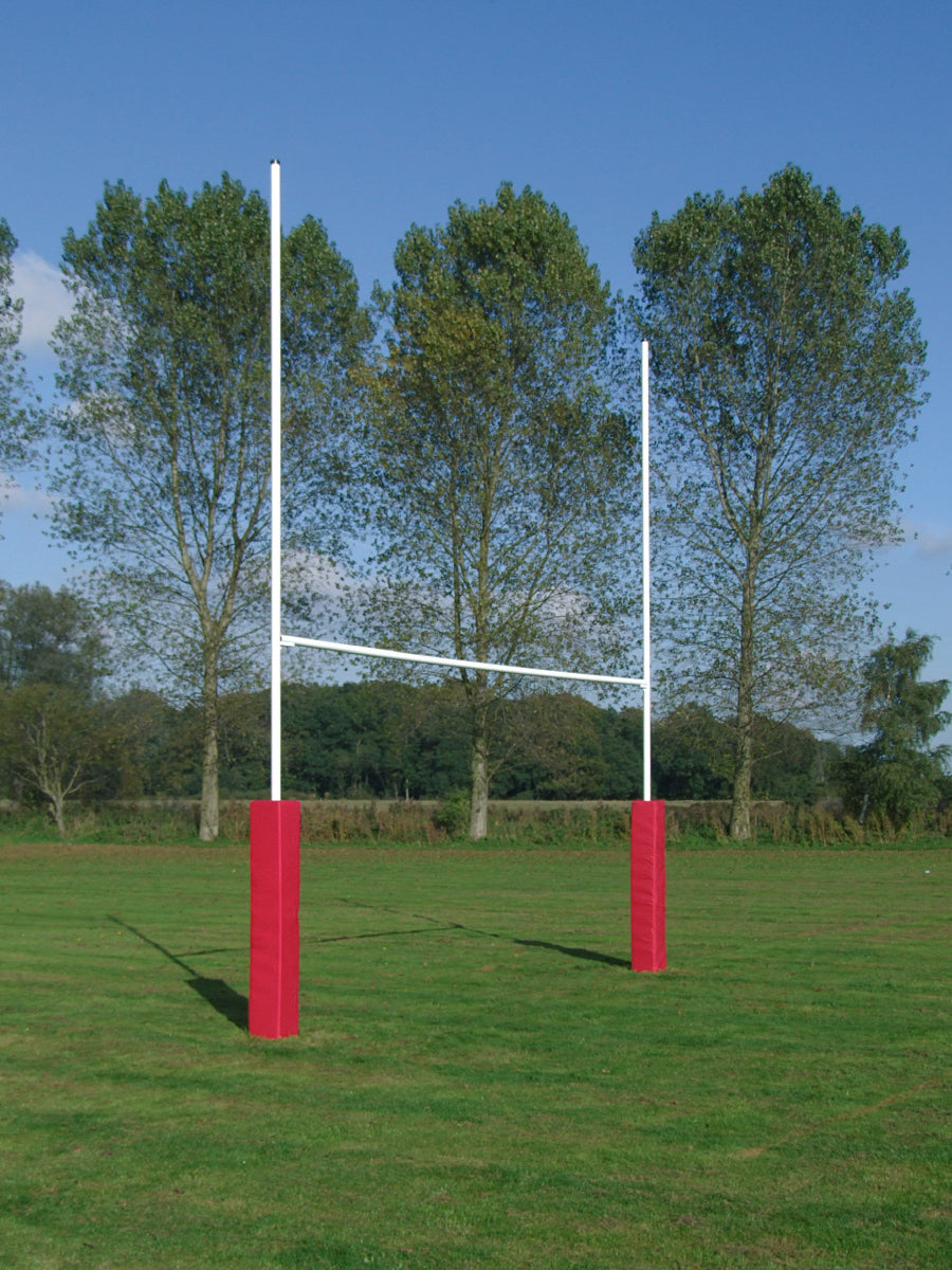 The Harrod Sport No 3 Steel Rugby Posts - 6M Socketed, featuring red padding, are set up on a grassy field among tall trees beneath a clear blue sky.