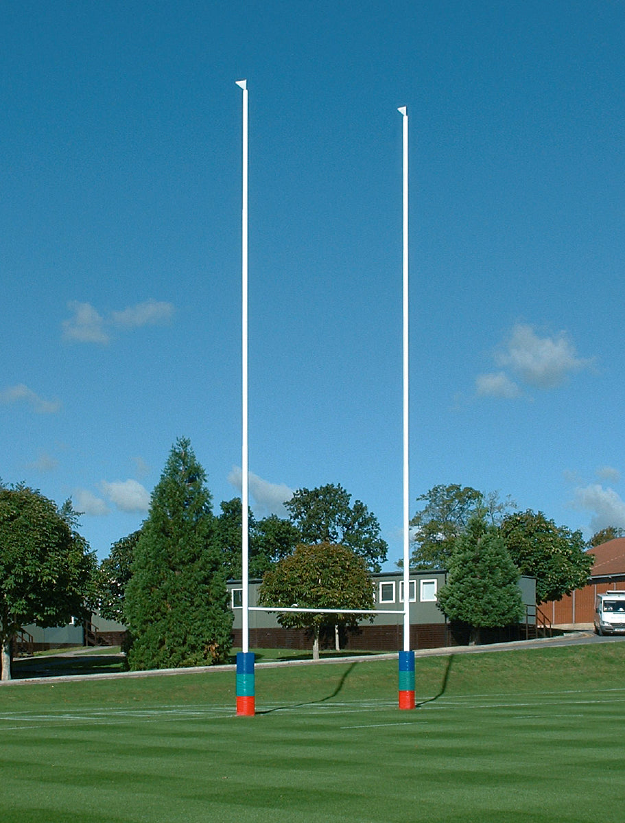 Harrod Sport Millennium Rugby Posts - 13.5M Hinged, with tall white aluminium uprights, a horizontal crossbar, and colorful base padding, stand on a grassy field bordered by trees and buildings beneath a clear blue sky.