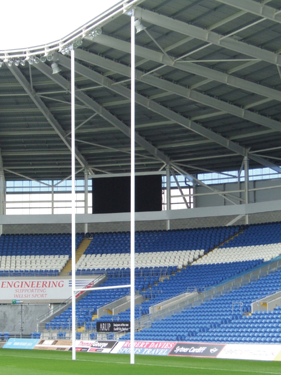Harrod Sport Aluminium Rugby Posts - 13M Hinged stand on a green field in a stadium featuring blue and white seats, advertising boards, and a partially covered roof.