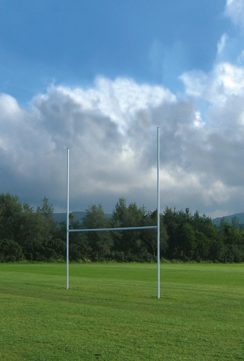 The Harrod Sport 5M Aluminium Mini Rugby Posts stand on a grassy field beneath a partly cloudy sky, with trees and distant hills in the background.