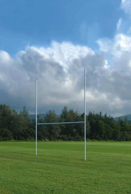The Harrod Sport 5M Aluminium Mini Rugby Posts stand on a grassy field beneath a partly cloudy sky, with trees and distant hills in the background.