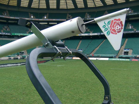 A close-up of a rugby corner flag with a red rose emblem, next to the Harrod Sport Hinged Post Assembly Roller on the grass field of an empty stadium with green seats in the background.