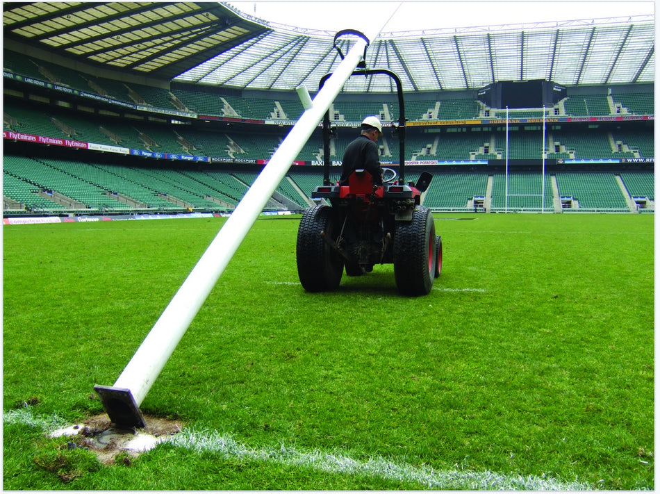 Using the Harrod Sport Hinged Post Assembly Roller, a person on a tractor with a roll bar installs or removes rugby posts on an empty stadium field with stands and seating visible in the background.