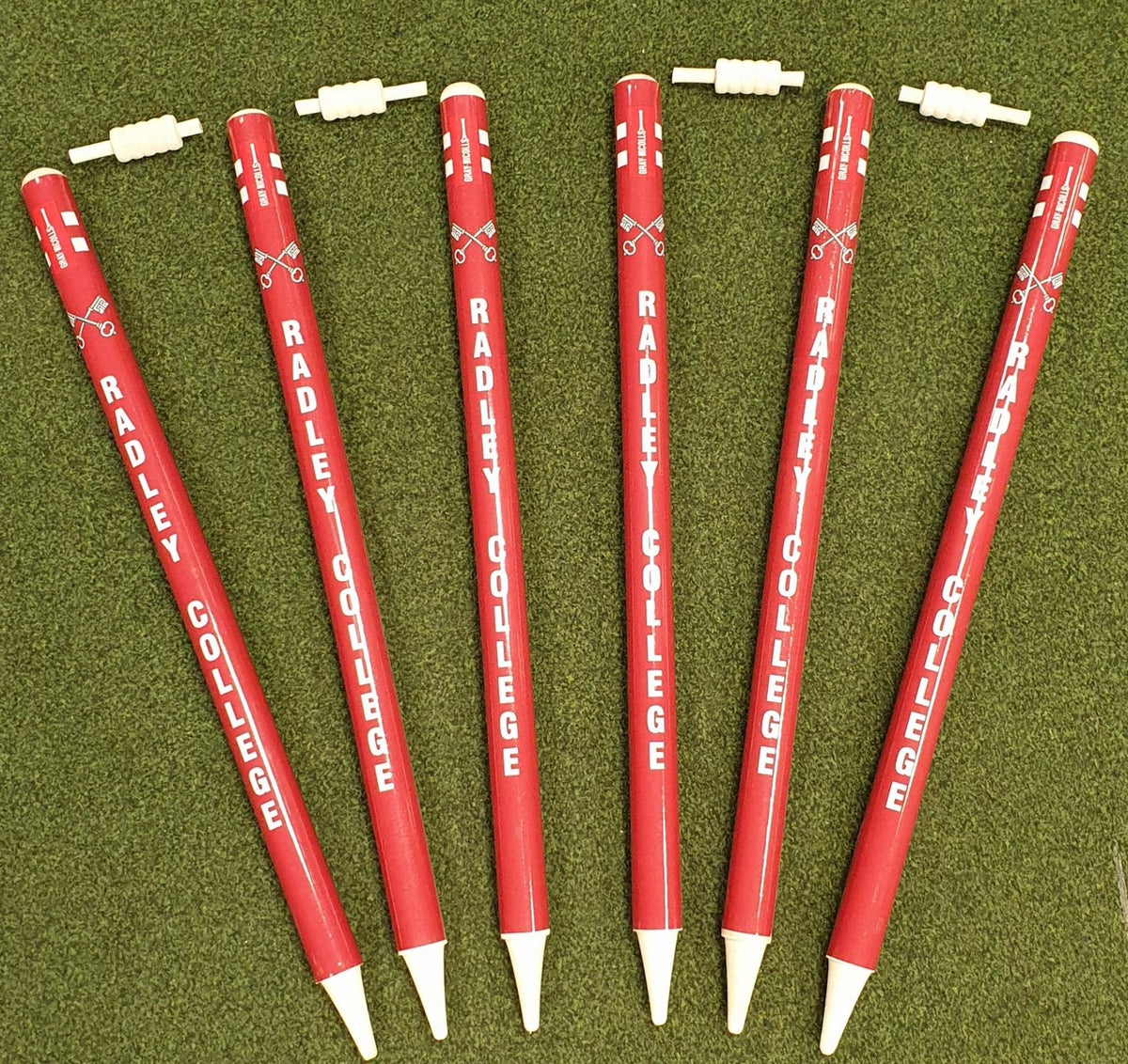 Six red Gray Nicolls Custom Printed Club Cricket Stumps with "RADLEY COLLEGE" in white are arranged fan-like on green grass, each featuring a white cap placed above or nearby, resembling personalised cricket stumps.