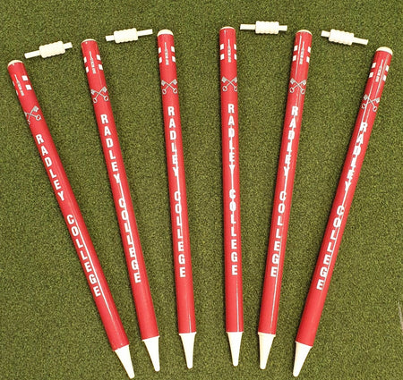 Six red Gray Nicolls Custom Printed Club Cricket Stumps with "RADLEY COLLEGE" in white are arranged fan-like on green grass, each featuring a white cap placed above or nearby, resembling personalised cricket stumps.