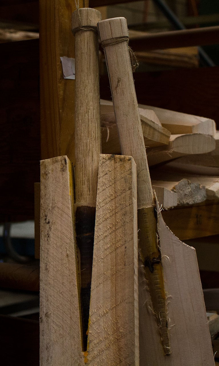 Two unfinished wooden cricket bats with rough handles stand in a holder amid wood shavings, illustrating the Cricket Bat Rehandle process by Martin Berrill Sports in a bustling workshop setting.