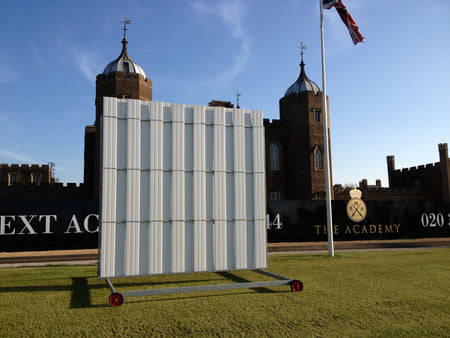 A large, movable Stuart Canvas Polypropylene Sightscreen with a metal frame and white panels stands on a grassy field before a historic brick building with domed towers and a flagpole. A sign for The Academy is visible in the background.