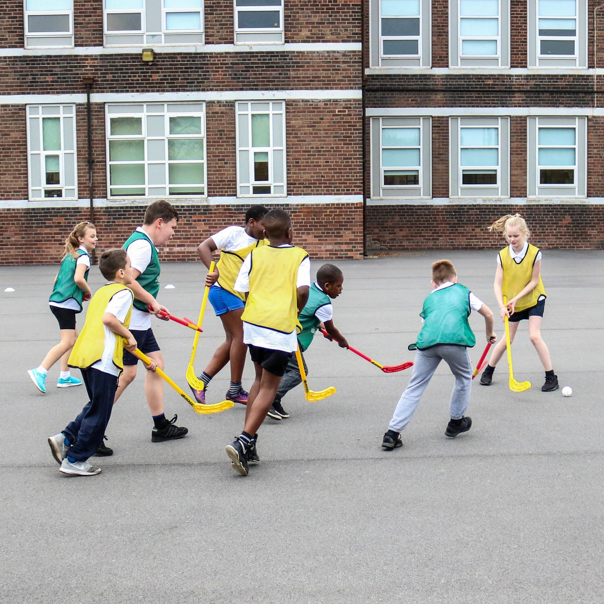 Children in yellow and green pinnies play with the Eurohoc Floorball Junior Set by Eurohoc, using plastic sticks on a paved schoolyard to build hand-eye coordination near a brick building with large windows.