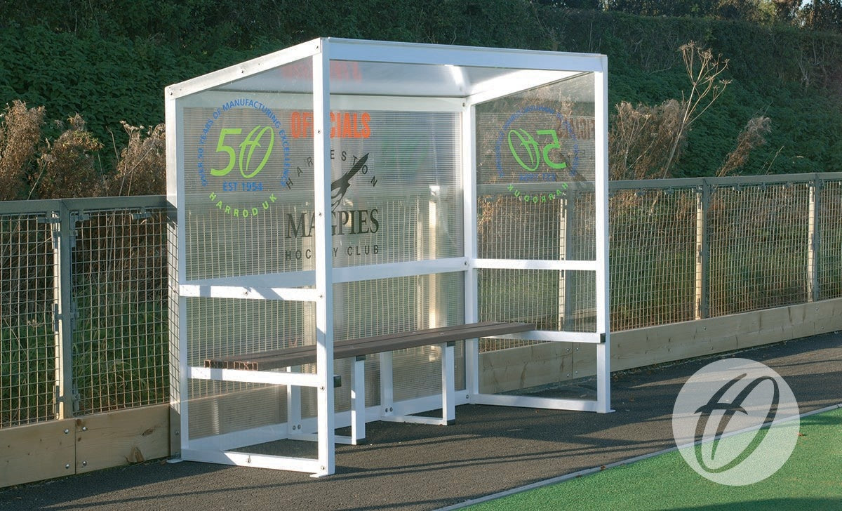 A Harrod Sport Aluminium Team Shelter - 2.5M Fixed with clear polycarbonate panels and a wooden bench is positioned beside a fenced sports field, featuring sponsor logos and greenery in the background.