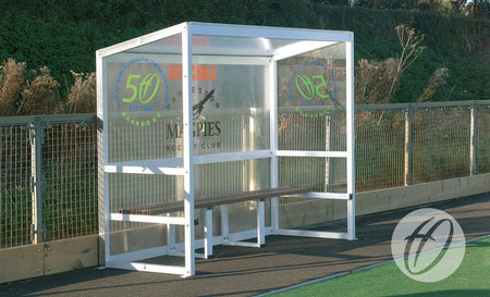 A Harrod Sport Aluminium Team Shelter - 2.5M Fixed with clear polycarbonate panels and a wooden bench is positioned beside a fenced sports field, featuring sponsor logos and greenery in the background.