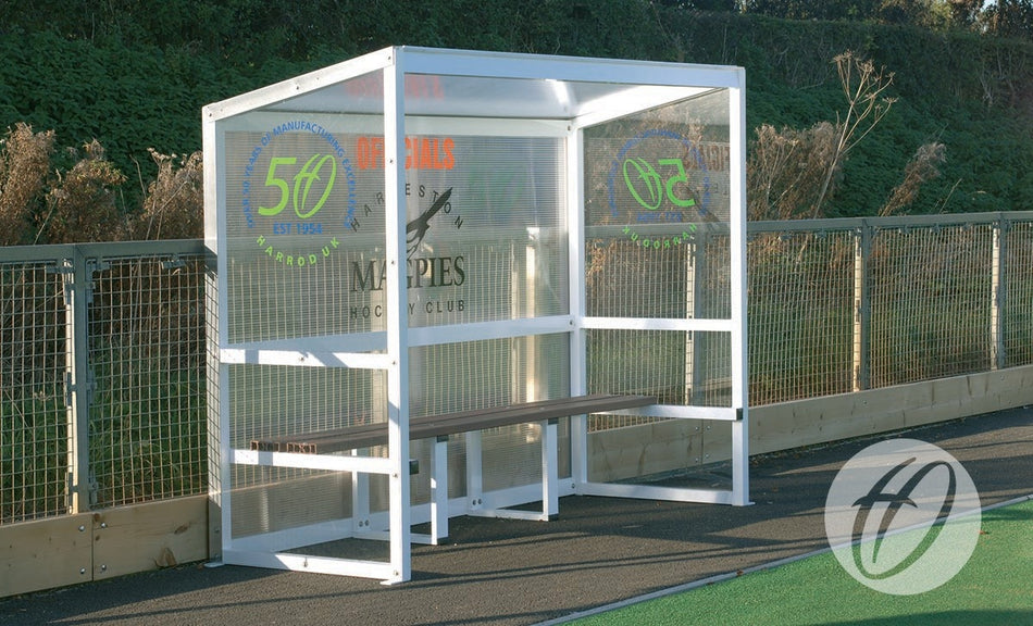 A Harrod Sport Aluminium Team Shelter - 2.5M Fixed with clear polycarbonate panels and a wooden bench is positioned beside a fenced sports field, featuring sponsor logos and greenery in the background.