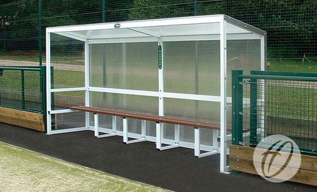 The Harrod Sport Aluminium Team Shelter - 4M Fixed features a metal frame, clear polycarbonate panels, wooden bench seating, and a covered roof for protection, ideal for placement beside fenced playing fields.