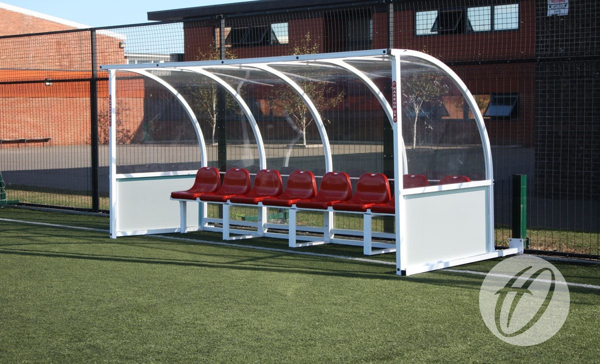 A Harrod Sport Premier Team Shelter - 4M Red Seats Fixed is floor mounted on the edge of an artificial turf field, providing seating for 8 beneath its clear curved roof, with buildings and fencing visible in the background.