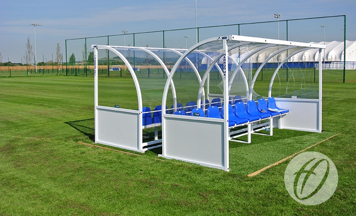 A Harrod Sport Premier Team Shelter - 4M Blue Seats Fixed stands on an empty, well-kept soccer field under a clear sky, with white fencing and sports facilities visible in the background.