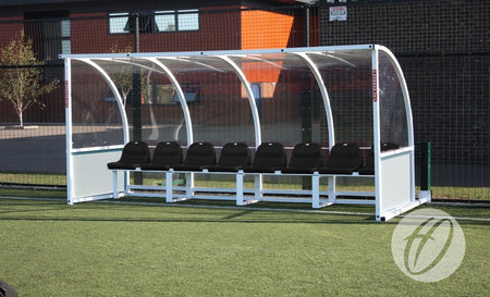 The Harrod Sport Premier Team Shelter- 4M features a transparent curved roof with eight fixed black seats, installed on artificial turf next to a black metal fence and a building in the background.