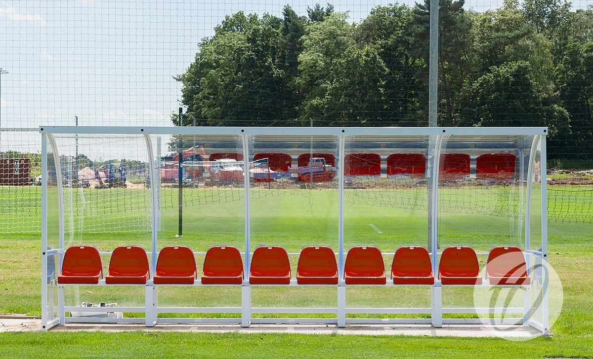 A Harrod Sport Premier Team Shelter - 5M with fixed red seats stands beside a green soccer field, sheltered by a transparent roof, with a goalpost and trees visible in the background.