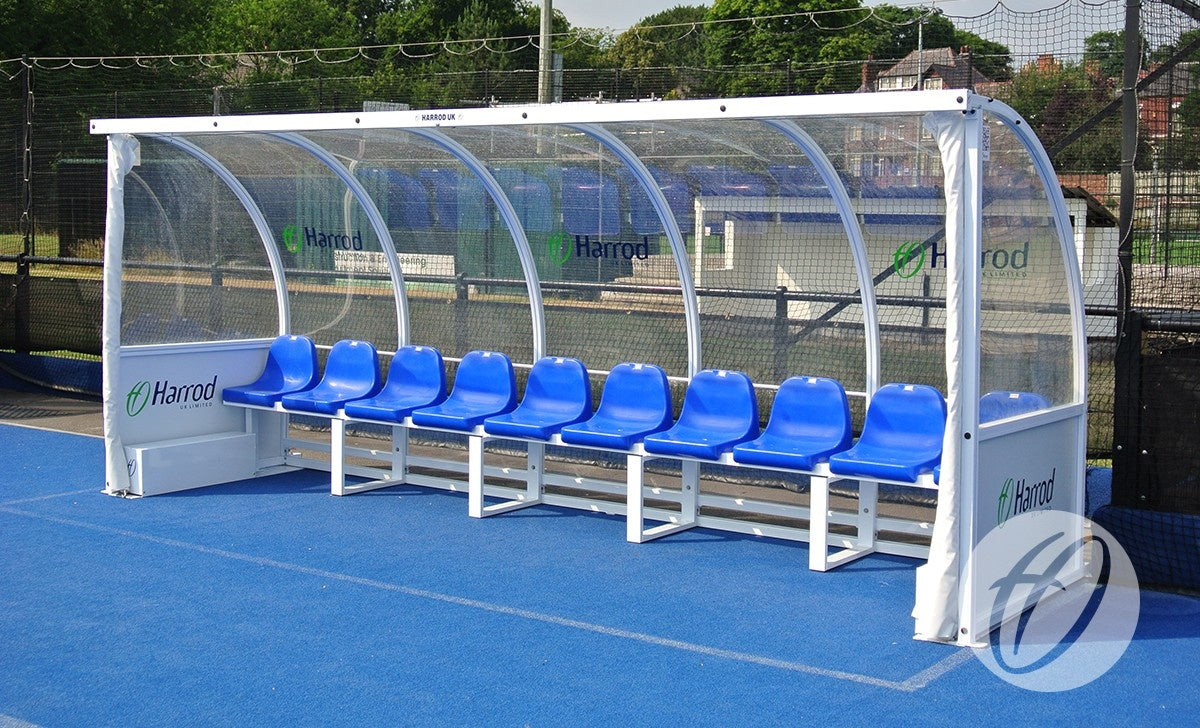 A row of 10 blue seats forms the Harrod Sport Premier Team Shelter - 5M Blue Seats Fixed, featuring a transparent curved roof and floor fixing, ideal for teams. The panels display the Harrod Sport logo.