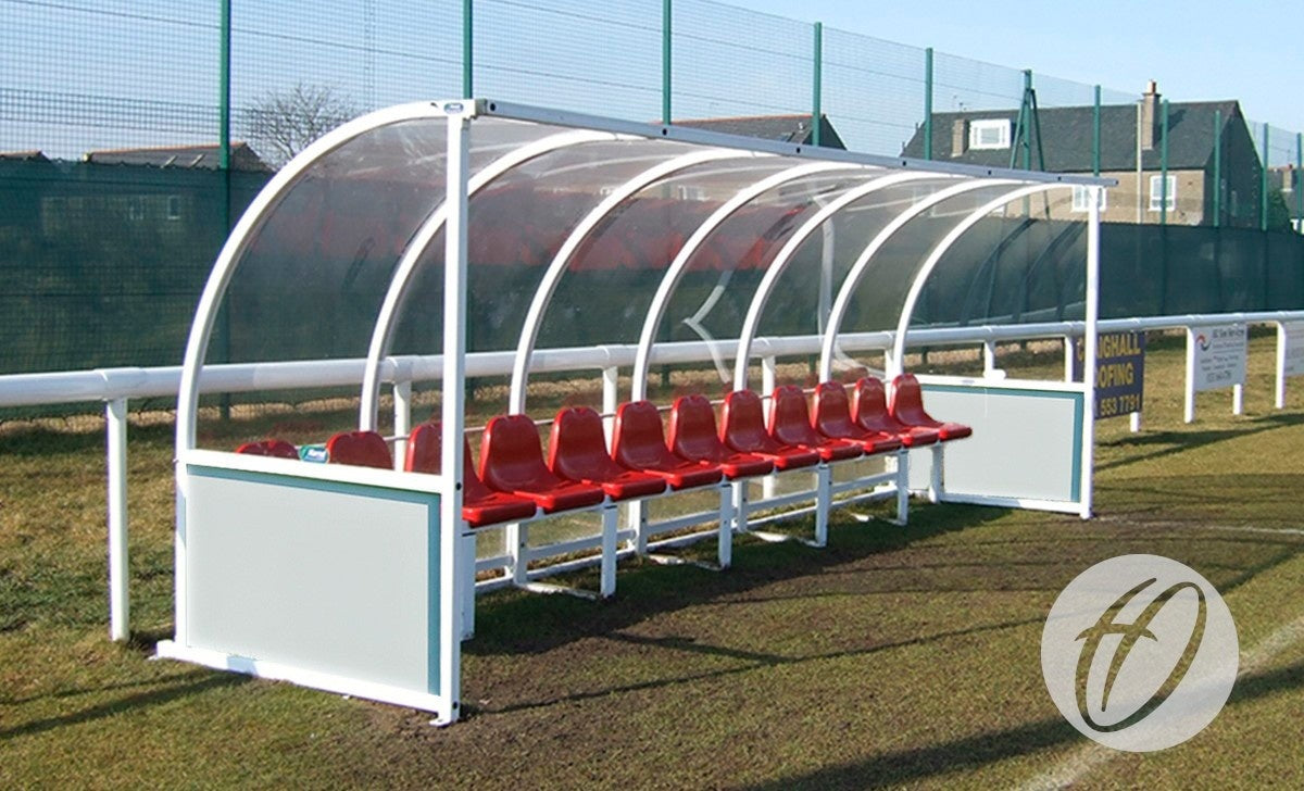 The Harrod Sport Premier Team Shelter - 6M Red Seats Fixed features red plastic seats and a transparent roof, floor-fixed beside a grassy field near fencing and buildings, providing durable comfort and protection for players.