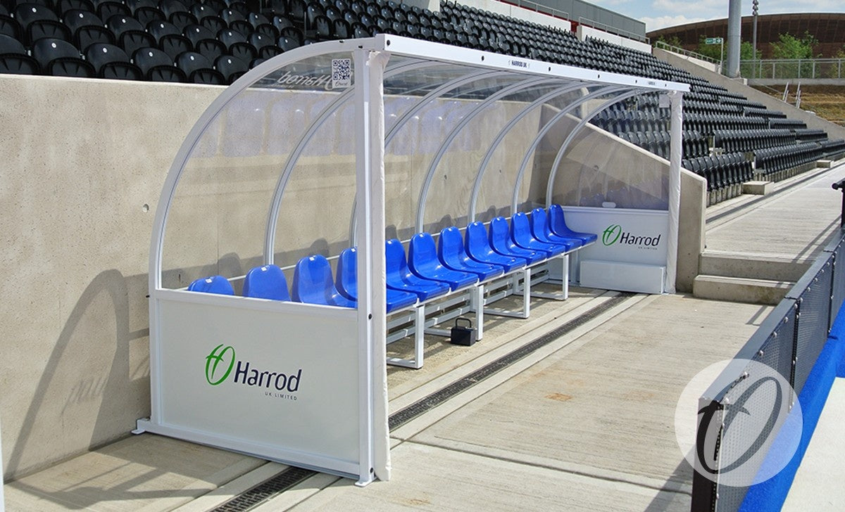 A row of blue seats sits beneath a transparent, curved Harrod Sport Premier Team Shelter (6m, 12 seats floor-fixed), beside a sports field and in front of empty stadium bleachers.