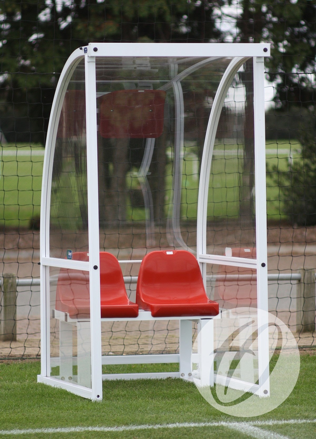 The Harrod Sport Premier Officials Shelter - 1M Red Seats Fixed is a compact aluminium shelter with a clear curved roof and two red seats, floor fixed on grass beside a sports field net, with trees in the background.
