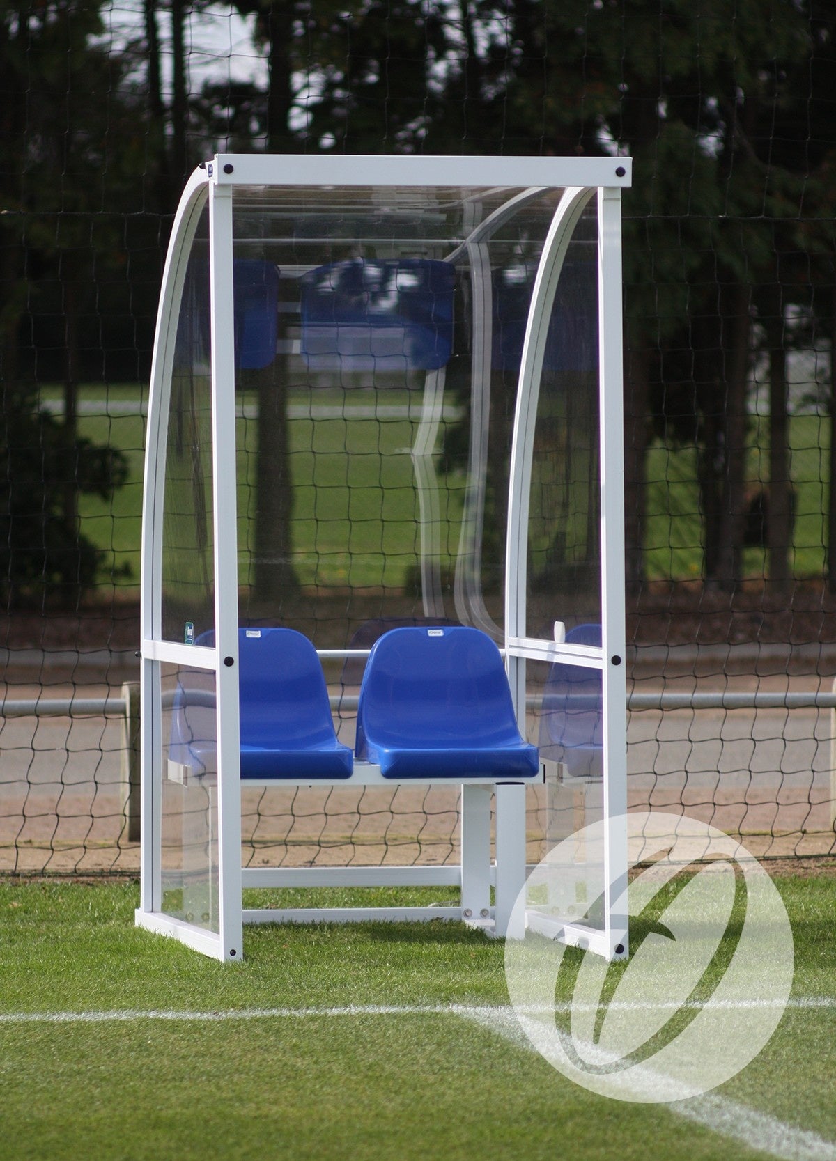 A Harrod Sport Premier Officials Shelter - 1M Blue Seats Fixed with a curved roof is positioned at the edge of a grassy football field. Two blue seats are inside, with trees and a netted fence visible in the background.