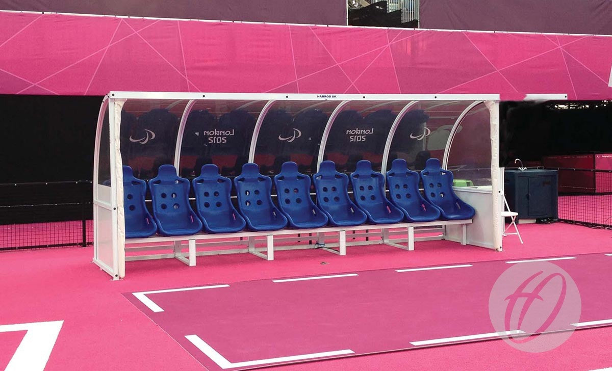 The Harrod Sport Superior Team Shelter - 6M Blue 10 Seats Fixed features clean blue bucket seats in an aluminium frame, providing sheltered seating for team players or coaches on a vibrant sports field.