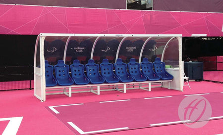 The Harrod Sport Superior Team Shelter - 6M Blue 10 Seats Fixed features clean blue bucket seats in an aluminium frame, providing sheltered seating for team players or coaches on a vibrant sports field.