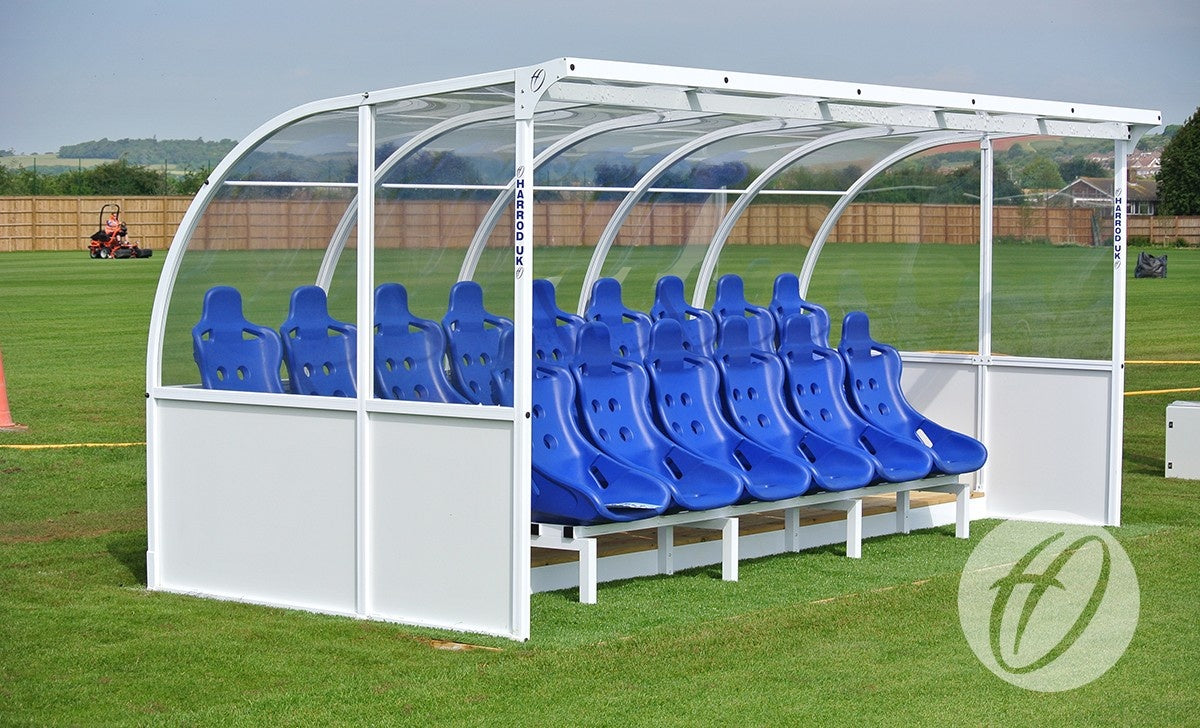 The Harrod Sport Elite Team Shelter - 5M features a row of blue seats under a clear, curved cover on the sports field, ideal for team substitutes. Fenced areas and someone on a red lawnmower are in the background.