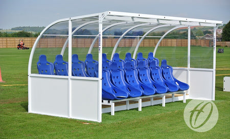 The Harrod Sport Elite Team Shelter - 5M features a row of blue seats under a clear, curved cover on the sports field, ideal for team substitutes. Fenced areas and someone on a red lawnmower are in the background.