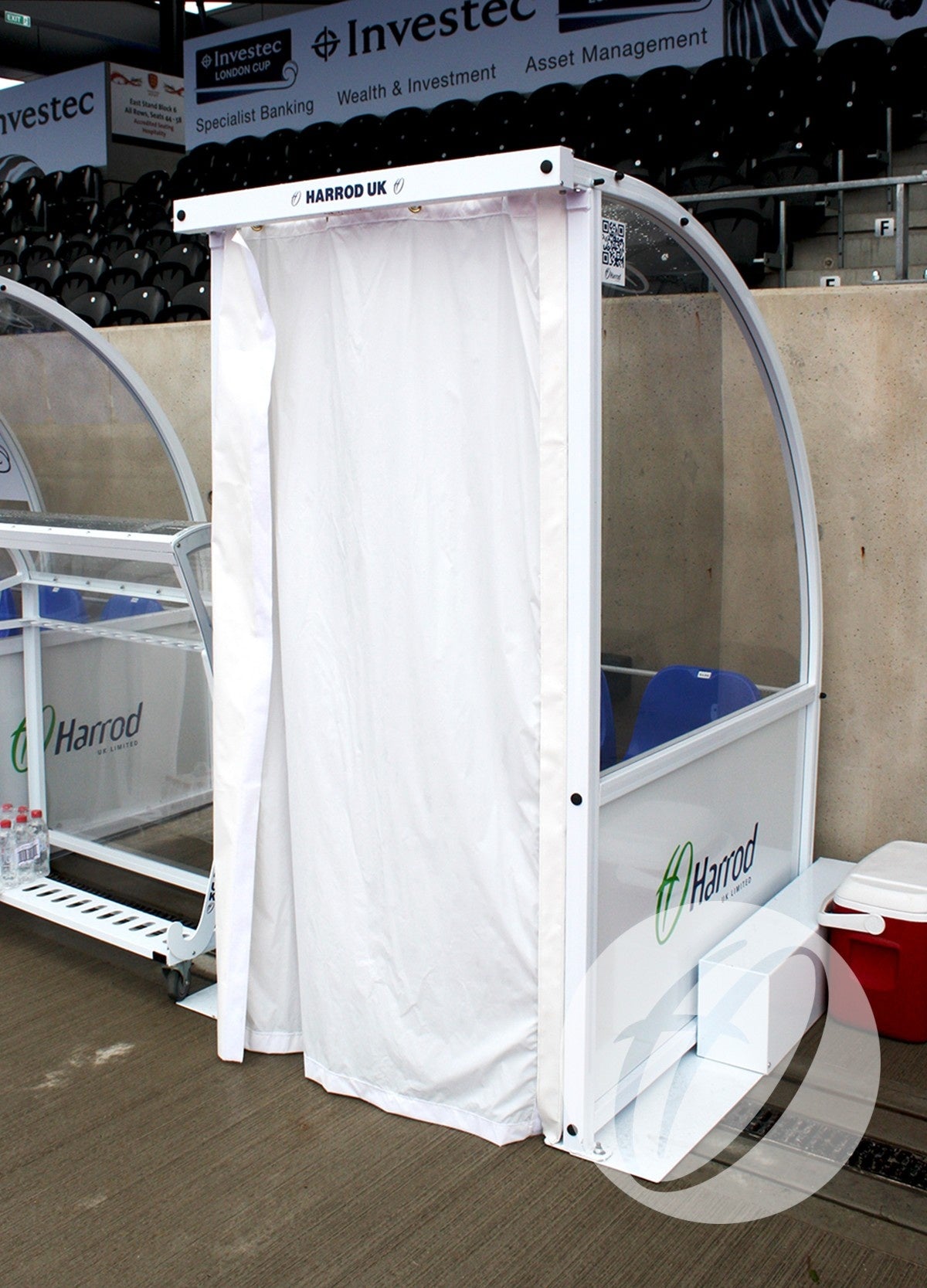 A white Curtain Pack For 1M Premier Team Shelters by Harrod Sport stands beside clear team benches at a stadium, featuring a waterproof privacy curtain on concrete near seating and advertising boards.