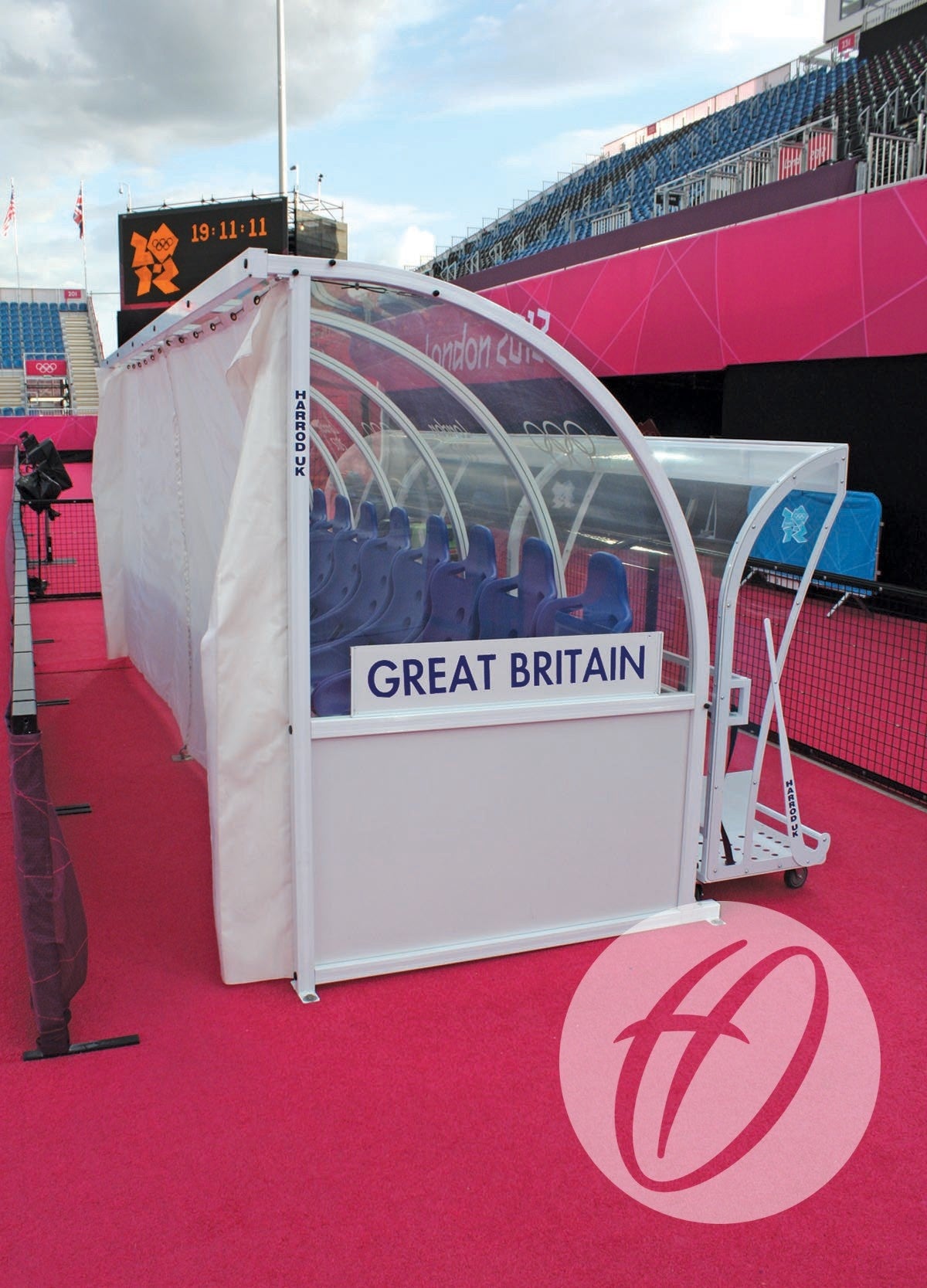 A Harrod Sport Curtain Pack for 4M Premier Team Shelters provides clear PVC weather protection to covered benches with Great Britain labels, seen on bright pink fields with blue seats and Olympics branding at London 2012.