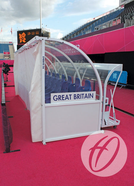 A Harrod Sport Curtain Pack for 4M Premier Team Shelters provides clear PVC weather protection to covered benches with Great Britain labels, seen on bright pink fields with blue seats and Olympics branding at London 2012.