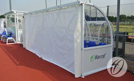A Harrod Sport Curtain Pack For 5M Premier Team Shelters covers a curved shelter on a red track surface beside a sports field, with the white PVC curtain drawn across the front to block the view and protect players outdoors.