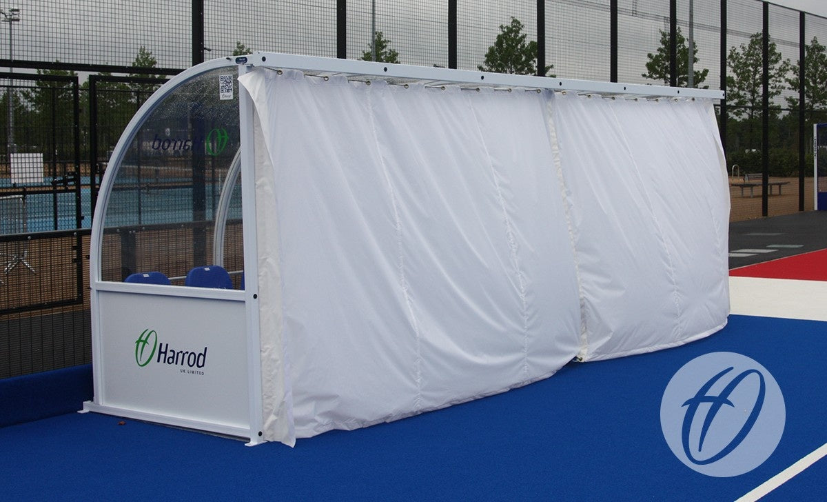 A white 6M Premier Team Shelter with a PVC Curtain Pack by Harrod Sport stands on a blue and red court near a fenced area with trees in the background.