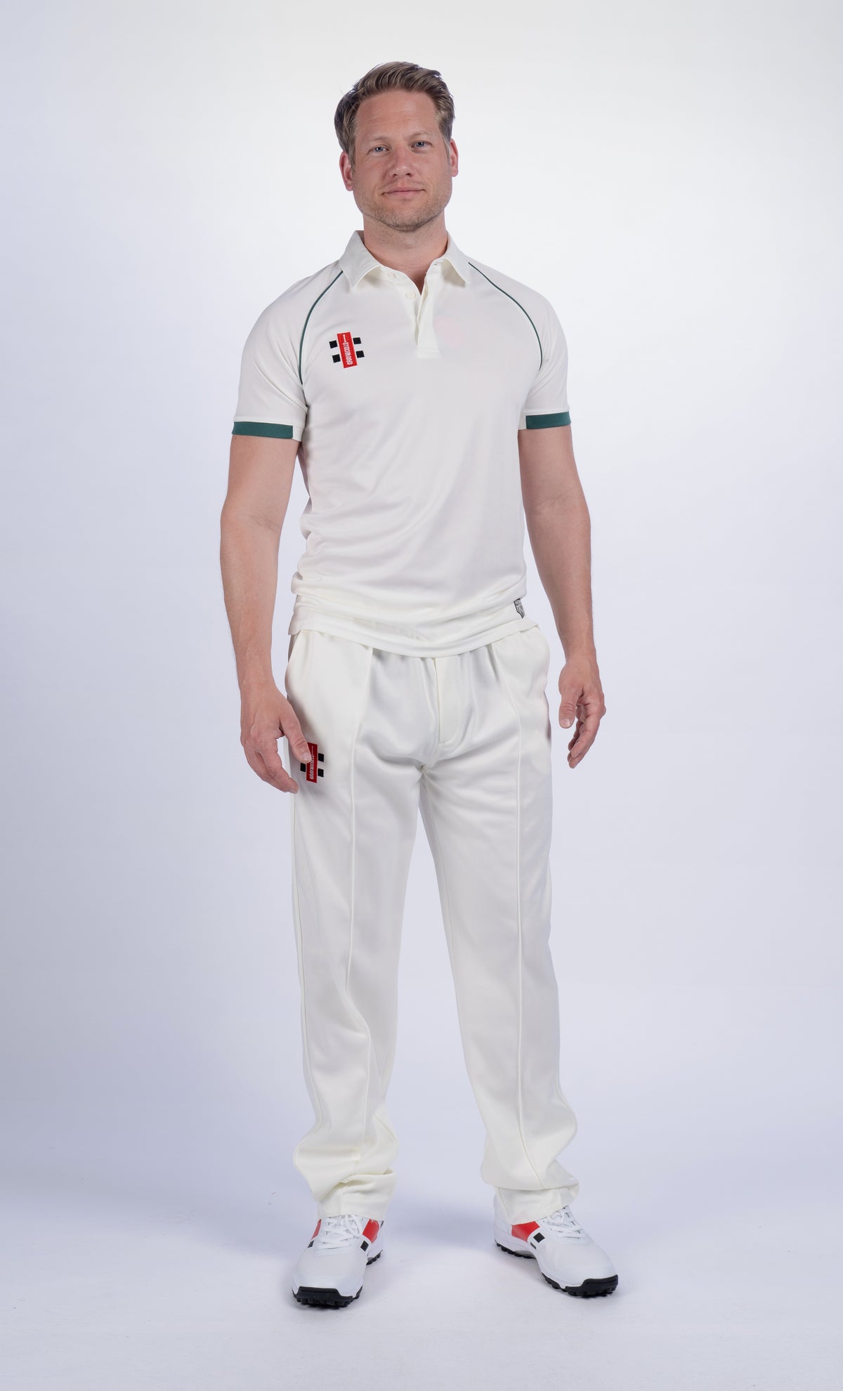 A man stands facing forward, wearing a Gray Nicolls Matrix V2 S/S Cricket Shirt—regular fit, white with green trim—and matching white sports shoes for moisture control, on a plain white background.