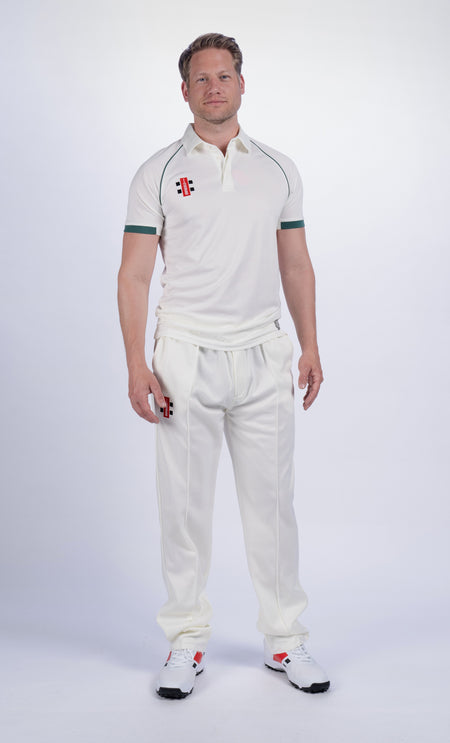 A man stands facing forward, wearing a Gray Nicolls Matrix V2 S/S Cricket Shirt—regular fit, white with green trim—and matching white sports shoes for moisture control, on a plain white background.