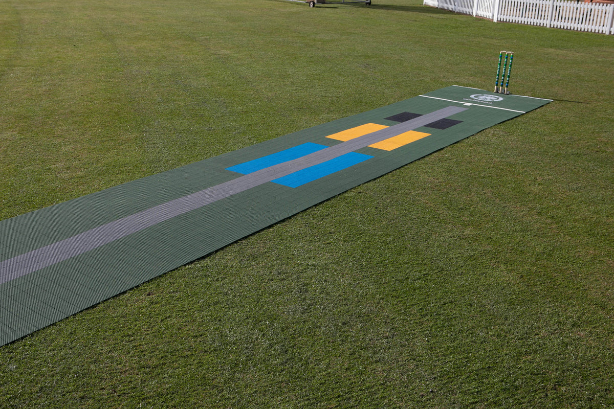 A Flicx UK 2G Flicx Skills & Eagle Eye Pitch mat, featuring blue, yellow, black, and gray markings, is set on grass with wickets at one end and a white fence behind—an ideal cricket coaching aid.
