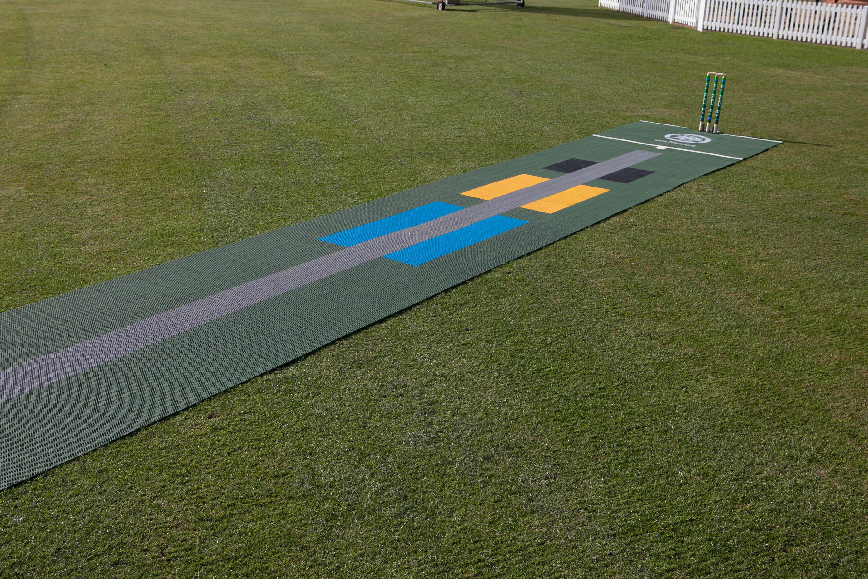 A Flicx UK 2G Flicx Skills & Eagle Eye Pitch mat, featuring blue, yellow, black, and gray markings, is set on grass with wickets at one end and a white fence behind—an ideal cricket coaching aid.