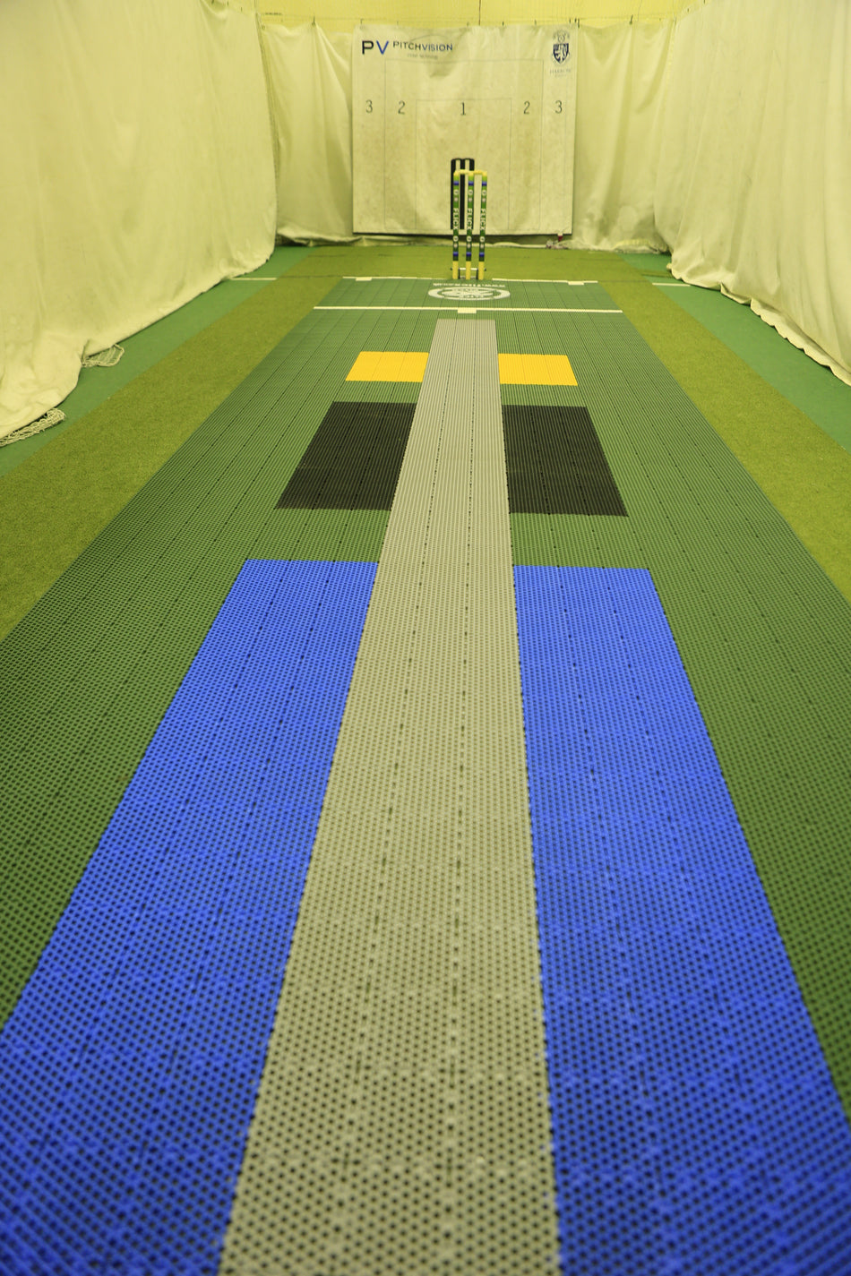 The Flicx UK 2G Flicx Skills & Eagle Eye Pitches aid cricket coaching with colored zones—blue, grey, black, yellow, green—leading to the stumps at the far end, complete with white curtains and a target board behind the stumps.