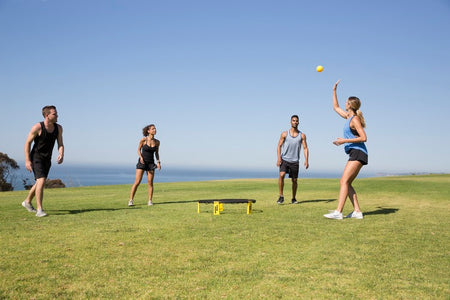 Four people play Spikeball near the ocean with a Kankakee Spikeball Standard Set, featuring an adjustable net. One woman tosses the yellow ball as the others get ready to join in under a clear blue sky.