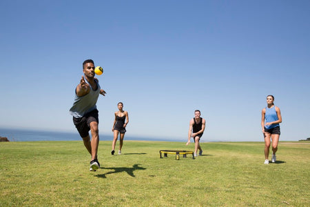Four people play with the Kankakee Spikeball Pro Set on grass near the ocean. One player lunges to hit the ball as three others watch, all under a clear, blue sky.