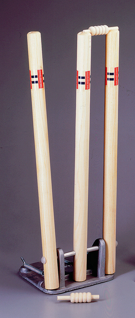The Gray Nicolls Senior Spring Return Stumps feature three wooden stumps with red and black accents set in a metal base, two bails on top, and one bail in front. Ideal for cricket training, shown against a plain gray background.