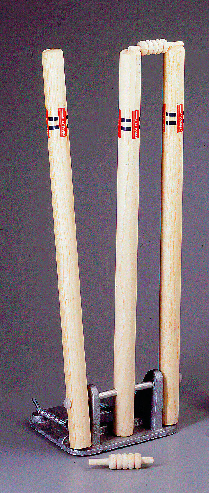 The Gray Nicolls Senior Spring Return Stumps feature three wooden stumps with red and black accents set in a metal base, two bails on top, and one bail in front. Ideal for cricket training, shown against a plain gray background.