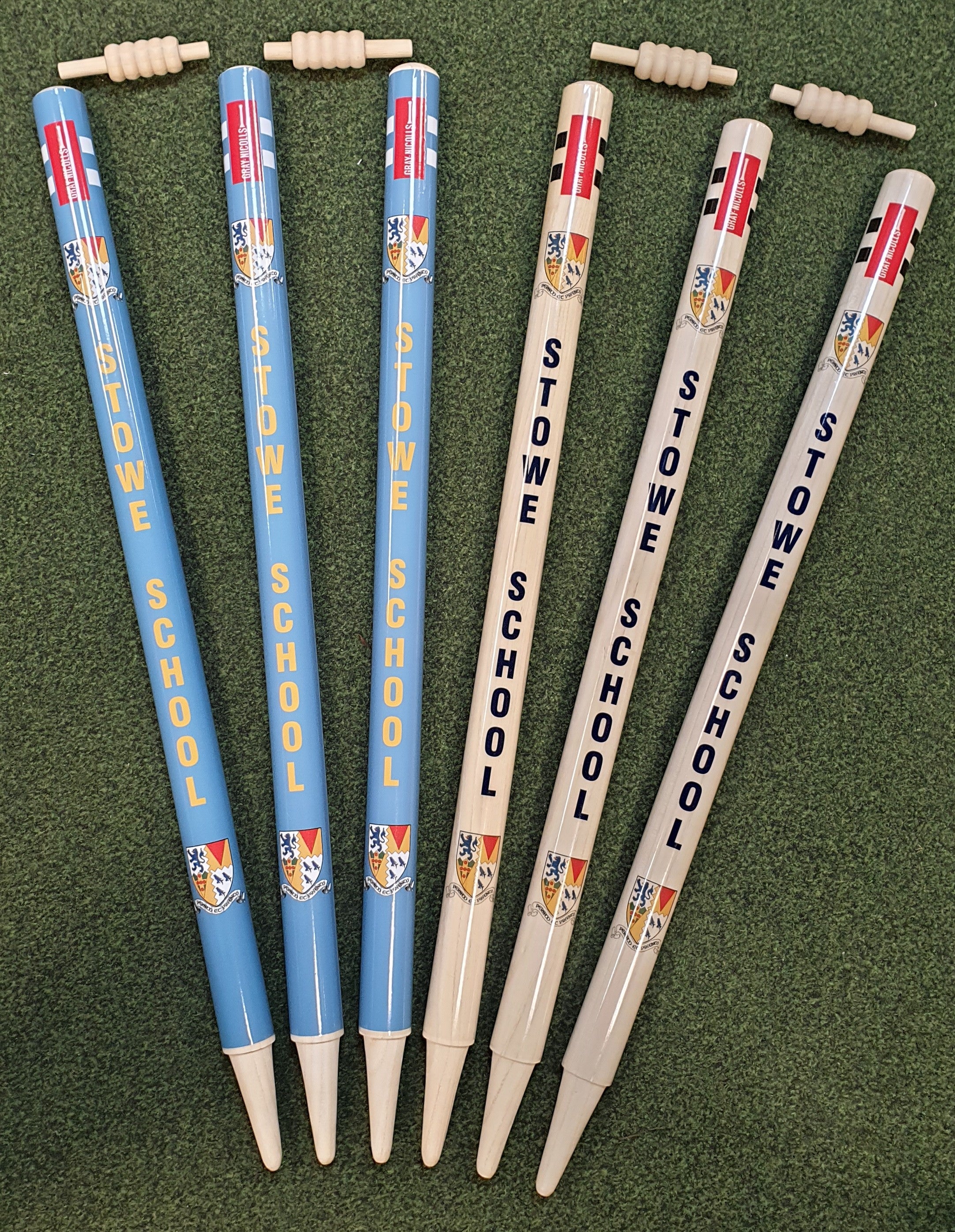 Six rounders bats, three blue and three beige, all labeled Stowe School with crests, are displayed on green grass beside Gray Nicolls Custom Printed Club Cricket Stumps for a smart sporting setup.