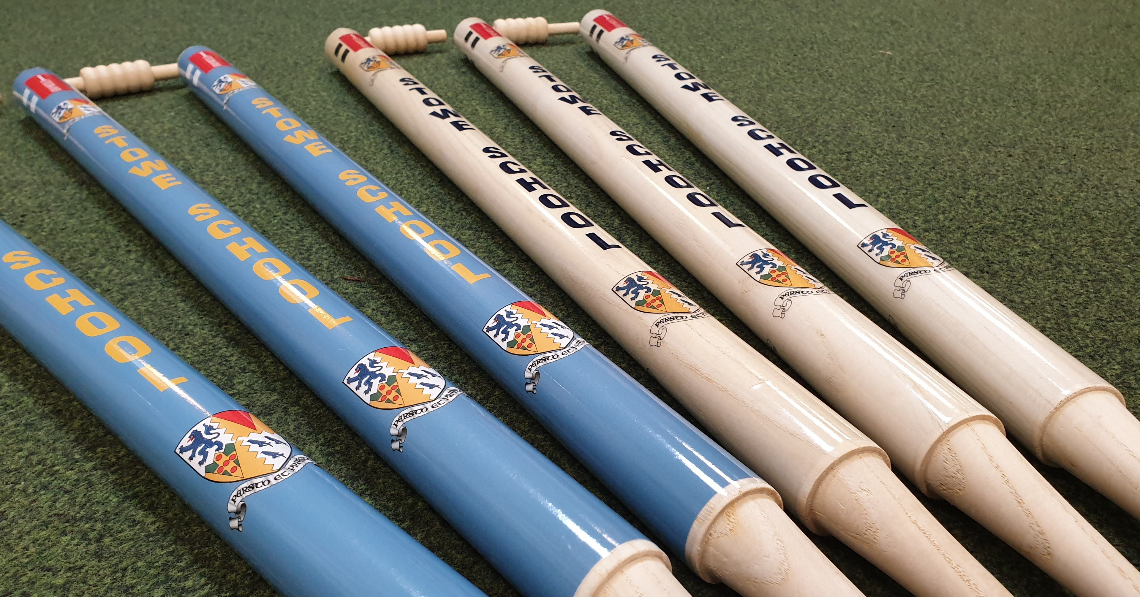 Five Gray Nicolls Custom Printed Club Cricket Stumps—three blue and two white—are displayed on a green field, each with HOUSE SCHOOL and a vibrant crest, ideal personalized cricket stumps for your club or school.