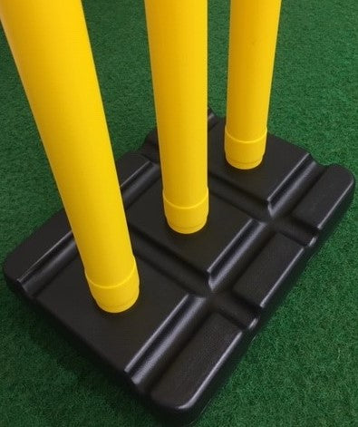 Close-up of three yellow stumps inserted into a Cricket Plastic Stump Base (Black) by Martin Berrill Sports, placed on green artificial turf.