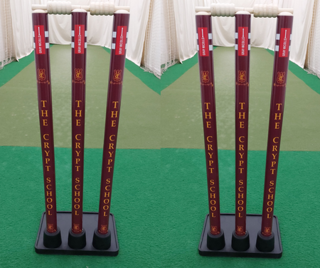 Two sets of maroon Gray Nicolls Custom Printed Match Cricket Stumps with Heavy Rubber Base, featuring "THE CRYPT SCHOOL" in yellow, stand on green artificial turf inside an indoor practice net.