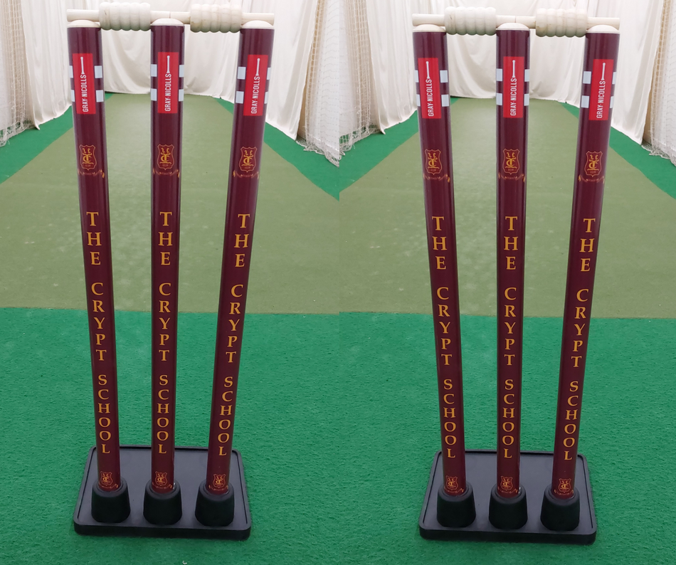 Two sets of maroon Gray Nicolls Custom Printed Match Cricket Stumps with Heavy Rubber Base, featuring "THE CRYPT SCHOOL" in yellow, stand on green artificial turf inside an indoor practice net.